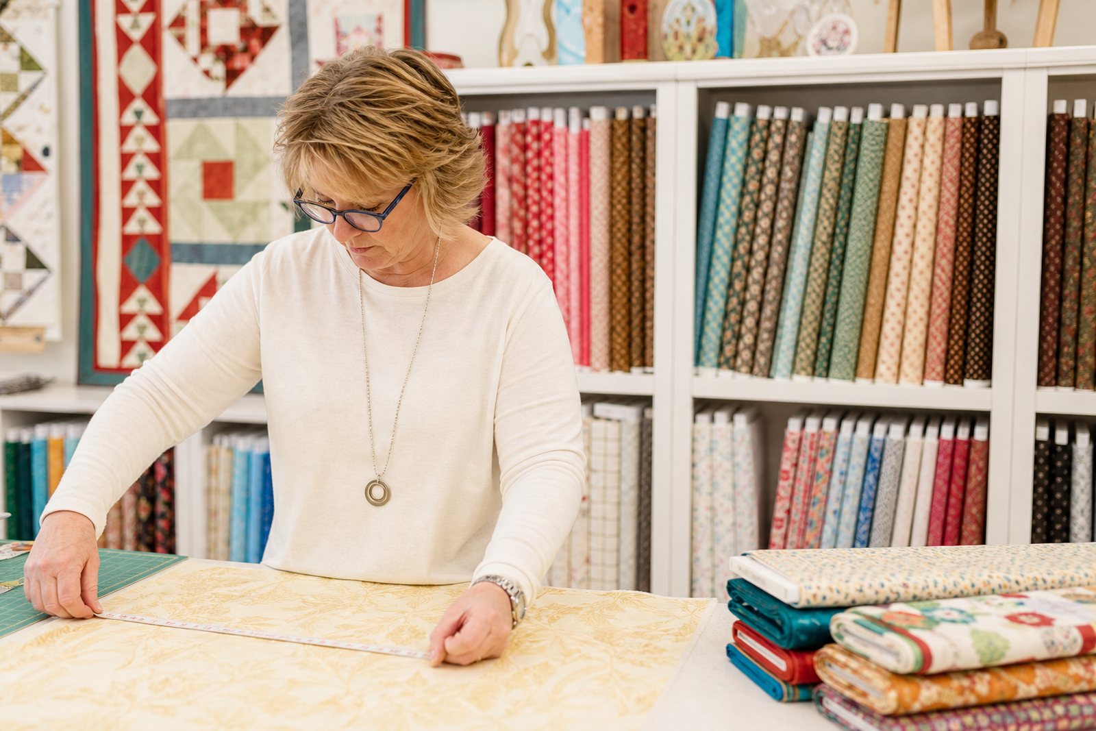 Quilt shop owner measuring a length of cotton at the cutting counter with shelves of colorful bolts behind her